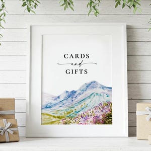 May include: A white frame with a watercolor painting of mountains and flowers. The text "CARDS and GIFTS" is printed in black at the top of the frame.