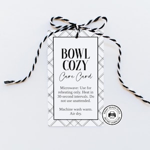 May include: A rectangular white tag with a gray plaid border and black text reading BOWL COZY Care Card, Microwave: Use for reheating only. Heat in 30-second intervals. Do not use unattended. Machine wash warm. Air dry. It is tied with black and white twine.
