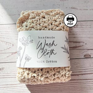 Op de afbeelding: Een beige gehaakt washandje van 100% katoen, omwikkeld met een witte papieren band met zwarte bloemmotieven en de tekst "handmade Wash Cloth 100% Cotton". Een rond icoon met een printer en de letters PDF is zichtbaar op de afbeelding.