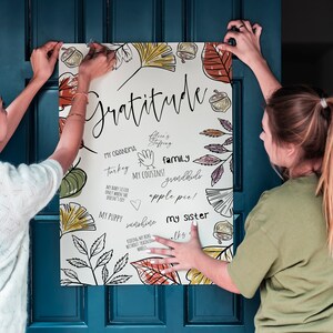 Peut inclure: Deux personnes accrochent une affiche blanche avec un motif d'automne sur une porte bleue. L'affiche porte le mot "Gratitude" &eacute;crit en cursive noire et pr&eacute;sente des dessins de feuilles, de glands et d'autres &eacute;l&eacute;ments d'automne. L'affiche comprend &eacute;galement une liste de choses pour lesquelles &ecirc;tre reconnaissant, telles que "Ma grand-m&egrave;re", "Dinde", "Mes cousins", "Famille", "Petits-enfants", "Tarte aux pommes", "Mon chiot", "Soleil", et "Ma s&oelig;ur".