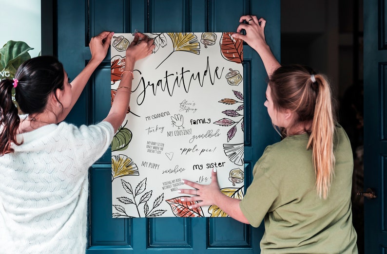 Peut inclure: Deux personnes accrochent une affiche blanche avec un motif d'automne sur une porte bleue. L'affiche porte le mot "Gratitude" &eacute;crit en cursive noire et pr&eacute;sente des dessins de feuilles, de glands et d'autres &eacute;l&eacute;ments d'automne. L'affiche comprend &eacute;galement une liste de choses pour lesquelles &ecirc;tre reconnaissant, telles que "Ma grand-m&egrave;re", "Dinde", "Mes cousins", "Famille", "Petits-enfants", "Tarte aux pommes", "Mon chiot", "Soleil", et "Ma s&oelig;ur".