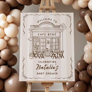 May include: A sign on an easel with the words "Welcome to Cafe Bebe" and "Celebrating Natalie's Baby Shower." The sign has a drawing of a cafe with tables and chairs. The background is striped, and there are balloons in shades of brown and cream.