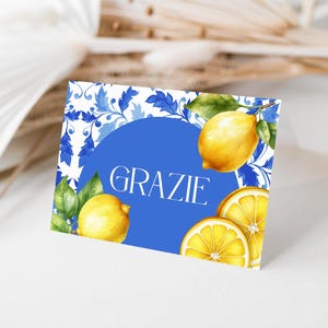 May include: A blue and white card with a blue circle in the center that says "GRAZIE" in white text. The card is decorated with lemons and leafy vines.