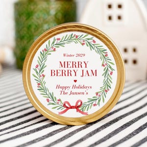 May include: A gold tin with a white label featuring a green wreath with red berries and a red ribbon. The label reads "Winter 2029 Merry Berry Jam Happy Holidays The Jansen's".
