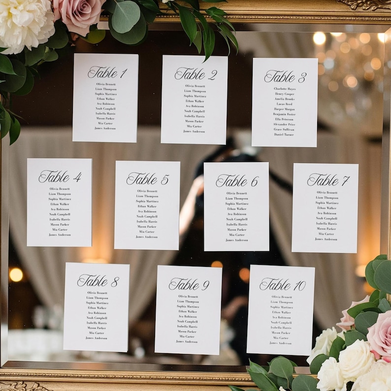 May include: A gold framed mirror displays ten white cards, each labeled with a table number and guest names in elegant script. Floral arrangements of pink and white roses and greenery frame the mirror, creating a wedding theme.