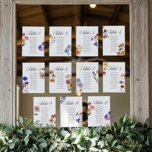 May include: Eleven white table cards with floral designs and guest names are displayed in a wooden framed mirror. Each card is labeled with a table number and features colorful watercolor flowers. The cards are arranged in a grid.