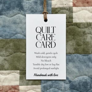 May include: A white rectangular tag with the words "QUILT CARE CARD" in black, hanging from a brown string. The tag includes care instructions: wash cold, gentle cycle, mild detergent only, no bleach, tumble dry low or lay flat, avoid prolonged sunlight. "Handmade with love" is written at the bottom.