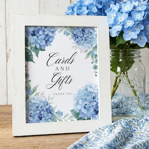Pode incluir: Uma moldura branca com uma placa que diz "Cartões e Presentes" com uma nota de agradecimento. A placa é decorada com hortênsias azuis e folhas verdes.