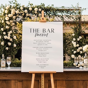 May include: A white bar menu sign on a wooden easel, with the words "THE BAR MENU" in elegant black script. The menu lists signature cocktails, wine, beer, and non-alcoholic options. The background features floral arrangements and glassware.