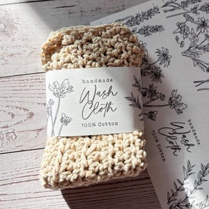 Op de afbeelding: Een beige gehaakt washandje van 100% katoen, omwikkeld met een witte papieren band met zwarte bloemmotieven en de tekst "handmade Wash Cloth 100% Cotton". Het washandje heeft een dikke, getextureerde structuur van geknoopt garen.