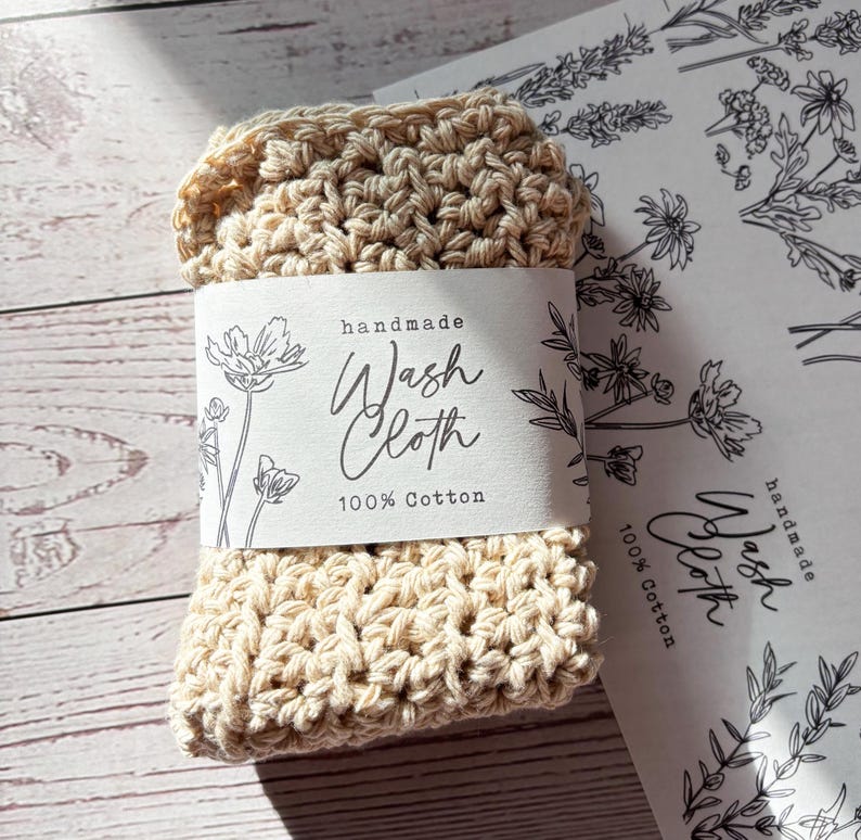Op de afbeelding: Een beige gehaakt washandje van 100% katoen, omwikkeld met een witte papieren band met zwarte bloemmotieven en de tekst "handmade Wash Cloth 100% Cotton". Het washandje heeft een dikke, getextureerde structuur van geknoopt garen.