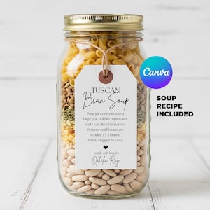 May include: A glass jar filled with ingredients for Tuscan Bean Soup, including pasta, beans, and herbs. A tag reads "Tuscan Bean Soup" with instructions and "Soup Recipe Included". The jar has a gold lid and is on a white wooden surface.