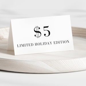 May include: A white card with the text "$5 LIMITED HOLIDAY EDITION" in black lettering. The card is propped up on a light-colored wooden tray with a circular shape.