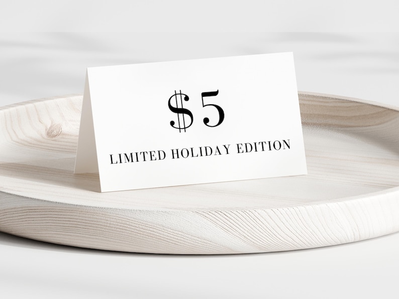 May include: A white card with the text "$5 LIMITED HOLIDAY EDITION" in black lettering. The card is propped up on a light-colored wooden tray with a circular shape.