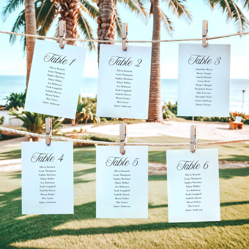 May include: Six white cards with guest names and table numbers, labeled "Table 1" through "Table 6", are clipped to a rope with wooden clothespins. The cards are displayed outdoors with palm trees and a beach in the background.