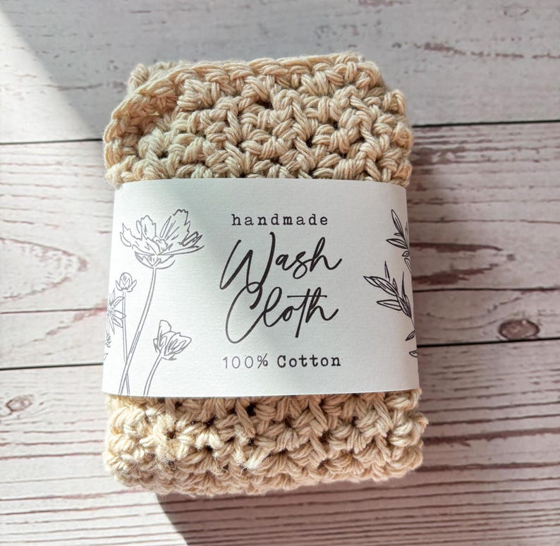 Op de afbeelding: Een beige gehaakt washandje van 100% katoen. Een witte papieren band zit om het midden, voorzien van zwarte florale lijntekeningen en de tekst: handmade Wash Cloth 100% Cotton. De textuur is dik, geknoopt en gelijkmatig van structuur.
