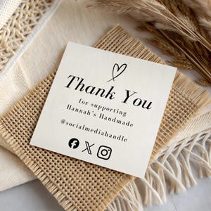 May include: A square, off-white card with the words "Thank You" in elegant black script, topped with a heart. The card reads "for supporting Hannah's Handmade" and includes social media icons. The card rests on a woven surface.