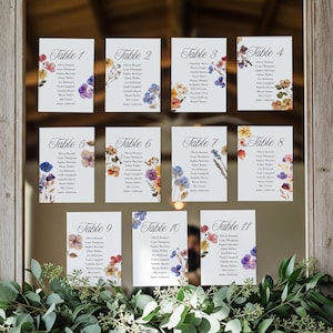 Könnte beinhalten: Elf weiße Tischkarten mit floralen Aquarelldesigns und Gästenamen. Jede Karte ist mit einer Tischnummer von 1 bis 11 versehen. Die Karten sind mit bunten Blumen in Lila-, Gelb- und Blautönen verziert.