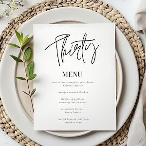 Op de afbeelding: Een witte menukaart met het woord "Thirty" in elegant zwart schrift. Het menu bevat gerechten zoals geroosterde biet, dragon gebraden kip en karnemelkkoekjes. De kaart ligt op een wit bord, met een geweven placemat en groen.