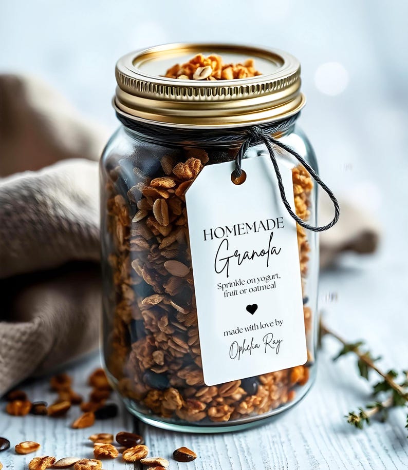 Op de afbeelding: Een glazen pot gevuld met zelfgemaakte granola, met een wit label waarop staat "HOMEMADE Granola Sprinkle on yogurt, fruit or oatmeal made with love by Ophelia Ray".