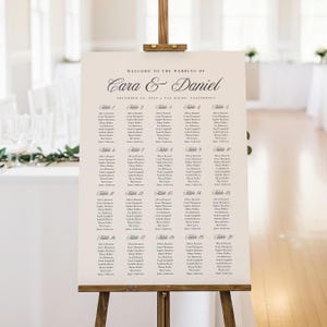 May include: A wedding seating chart on a wooden easel. The sign is cream-colored with elegant script that reads "Welcome to the Wedding of Cara & Daniel." The chart lists guest names under table numbers 1-20.