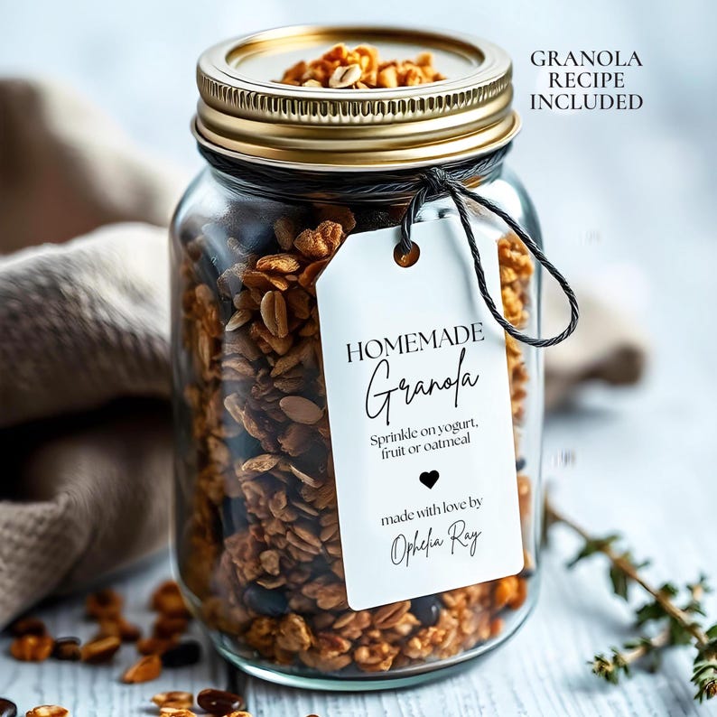 Op de afbeelding: Een glazen pot gevuld met zelfgemaakte granola, vastgebonden met een zwart lint en een wit label met de tekst "HOMEMADE Granola Sprinkle on yogurt, fruit or oatmeal made with love by Ophelia Ray".