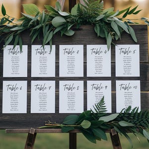 May include: A wooden sign with ten white cards displaying table numbers and guest names in black script. The sign is decorated with green foliage. Perfect for a wedding or event.