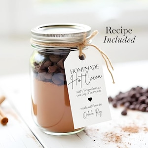 Op de afbeelding: Een glazen pot gevuld met ingredi&euml;nten voor zelfgemaakte warme chocolademelk, met een wit label dat om de pot is gebonden. Het label luidt "HOMEMADE Hot Cocoa" en bevat instructies voor het maken van de warme chocolademelk. Het label bevat ook de tekst "made with love by Ophelia Ray".