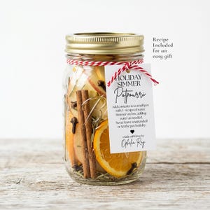 May include: A clear glass mason jar with a gold lid filled with dried orange slices, cinnamon sticks, and cloves. A white tag tied with red and white twine reads Holiday Simmer Potpourri and includes instructions and the name Ophelia Ray.