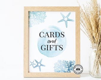 May include: A light wood frame containing a white sign with the text CARDS and GIFTS in black font. The sign features a central light blue watercolor circle, light blue starfish illustrations, and blue coral patterns on a white background.