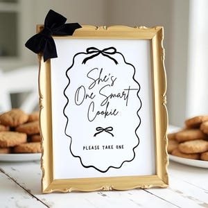 May include: A gold-colored frame with a white sign that says "She's One Smart Cookie" with a black bow on top. The sign is on a white table with a plate of cookies.