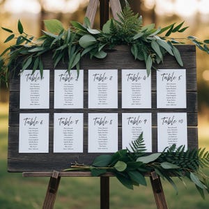 May include: A wooden easel displaying a seating chart for a wedding or event. The chart features ten tables, each labeled with a number and names. Greenery, including eucalyptus and ferns, decorates the top and bottom of the chart.