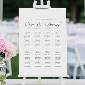May include: A white wedding seating chart on an easel, with elegant calligraphy announcing "Cara & Daniel." The chart lists guest names under table numbers. The wedding date and location are also displayed.