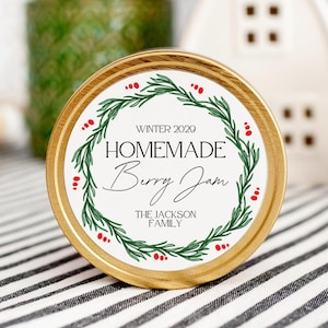 Pode incluir: Pote de metal dourado com um rótulo branco que apresenta uma grinalda verde com bagas vermelhas. O rótulo diz "Winter 2029 Homemade Berry Jam The Jackson Family".