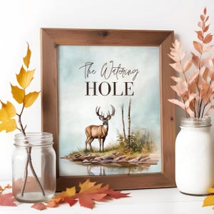 May include: A framed print with a watercolor illustration of a deer standing in a forest by a lake. The text "The Watering HOLE" is printed in brown at the top of the print.