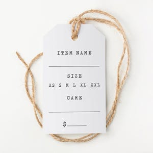Puede incluir: Una etiqueta blanca con las palabras "ITEM NAME", "SIZE" y "CARE" impresas en negro. Se enumeran las tallas XS, S, M, L, XL y XXL. Un símbolo de euro y una línea están en la parte inferior. La etiqueta está sujeta a un cordel natural.