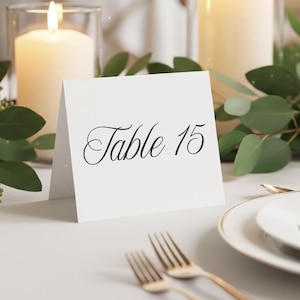 May include: A white table card with the text "Table 15" in elegant black script. The card is set on a table with gold forks, a white plate, and lit candles in the background, with greenery accents.