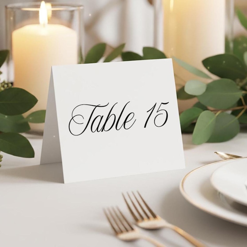 May include: A white table card with the text "Table 15" in elegant black script. The card is set on a table with gold forks, a white plate, and lit candles in the background, with greenery accents.