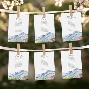 May include: Six table cards with watercolor mountain scenery and a list of names for each table. The cards are numbered one through six and are hanging from a white ribbon with clothespins.