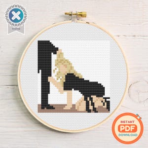 May include: A cross-stitch embroidery hoop with a black and white pixelated design of two figures. One figure is kneeling, and the other is standing. The embroidery is set against a white background. The hoop is made of wood and has a brass screw.