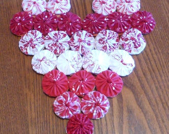 May include: A heart-shaped arrangement of red and white fabric rosettes. The rosettes are arranged in a pattern, with the larger rosettes in the center and the smaller rosettes on the edges.