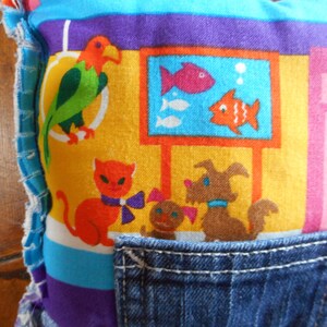 Op de afbeelding: Een kleurrijk kussen met een denim zak. Het kussen heeft een levendige print van een papegaai, vissen in een tank, een kat met een strik en een hond. Het ontwerp bevat gele, oranje, blauwe, paarse en roze tinten.