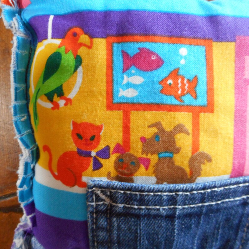 Op de afbeelding: Een kleurrijk kussen met een denim zak. Het kussen heeft een levendige print van een papegaai, vissen in een tank, een kat met een strik en een hond. Het ontwerp bevat gele, oranje, blauwe, paarse en roze tinten.