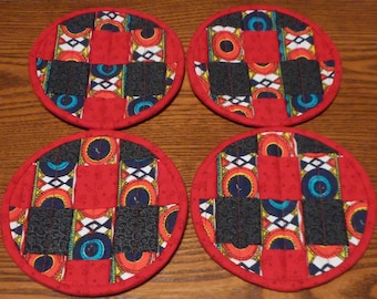 May include: Four round red fabric coasters with a patchwork design of black, white, yellow, blue, and red fabric. The coasters have a circular pattern of circles and stripes.