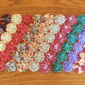 May include: A colorful table runner made of fabric rosettes in shades of pink, purple, green, red, and blue. The rosettes are arranged in a diagonal pattern.