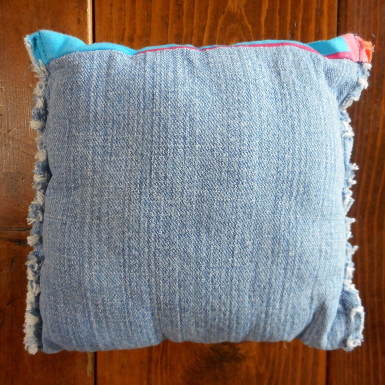 Op de afbeelding: Vierkantig denim kussen in een lichtblauwe tint. Het kussen heeft een gefranjerde rand en een contrasterende blauwe rand met roze en oranje accenten. Gemaakt van een zachte, getextureerde stof.