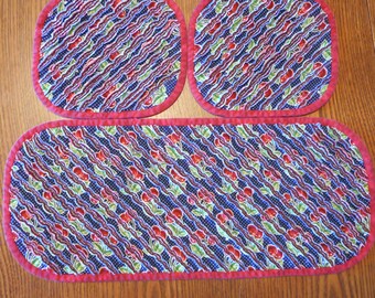 May include: A set of three quilted table mats with a red and blue cherry pattern. The mats are made of fabric and have a red border.