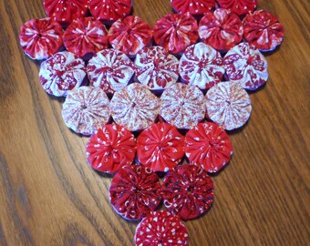 May include: A heart-shaped arrangement of red and white fabric rosettes. The rosettes are sewn together to create a heart shape. The rosettes are all different sizes and have a variety of patterns.