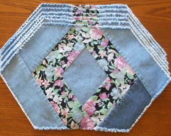 May include: A denim placemat with a floral fabric center. The placemat is made from multiple layers of denim fabric with frayed edges. The floral fabric is black with pink, white, and green flowers.