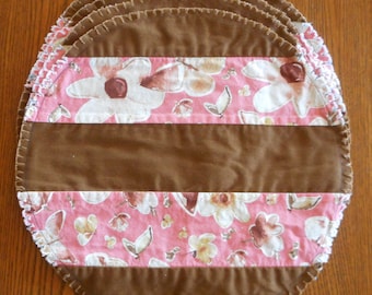 May include: A brown and pink fabric placemat with a floral pattern and brown trim. The placemat is made of multiple layers of fabric and has a scalloped edge.